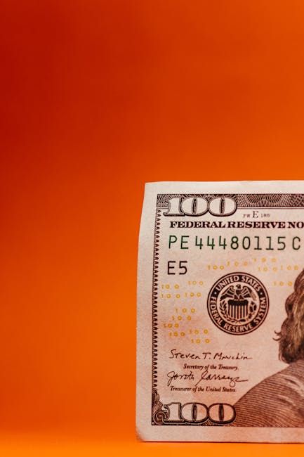 A close-up view of a transparent hourglass with black sand inside, positioned upright on a white surface. Behind the hourglass, there are several US dollar bills, including a twenty-dollar note, parti