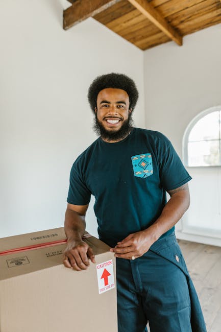 A professional mover with dark curly hair and a beard, standing inside a bright room with wooden ceiling beams and a large window, holding a large cardboard box labeled with a red caution arrow and te