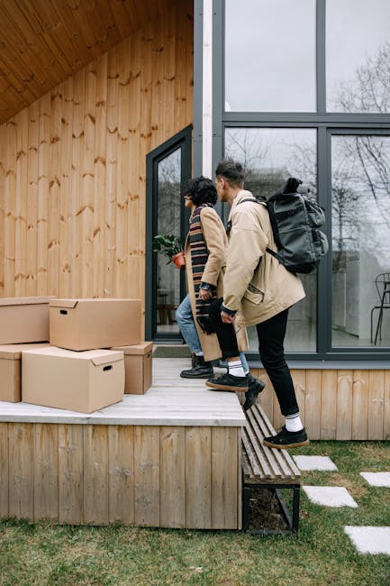 A woman with curly brown hair, wearing a beige fleece coat, blue jeans, and black shoes, is capturing a step in home relocation by carefully carrying a cardboard box with a potted plant on top down a