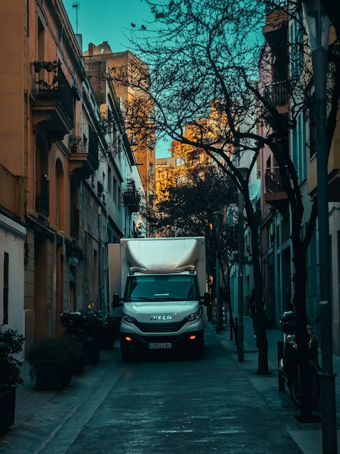 A white moving van parked on a city street in front of a row of shops, with the rear door open revealing a partially loaded interior containing cardboard boxes, plastic containers, and packing materia