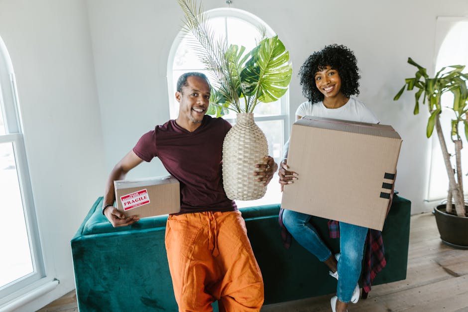 A man and woman indoors during a home relocation process, both holding cardboard boxes; the man is holding a smaller box with a red 'Fragile' label and an large beige ceramic vase with green tropical 